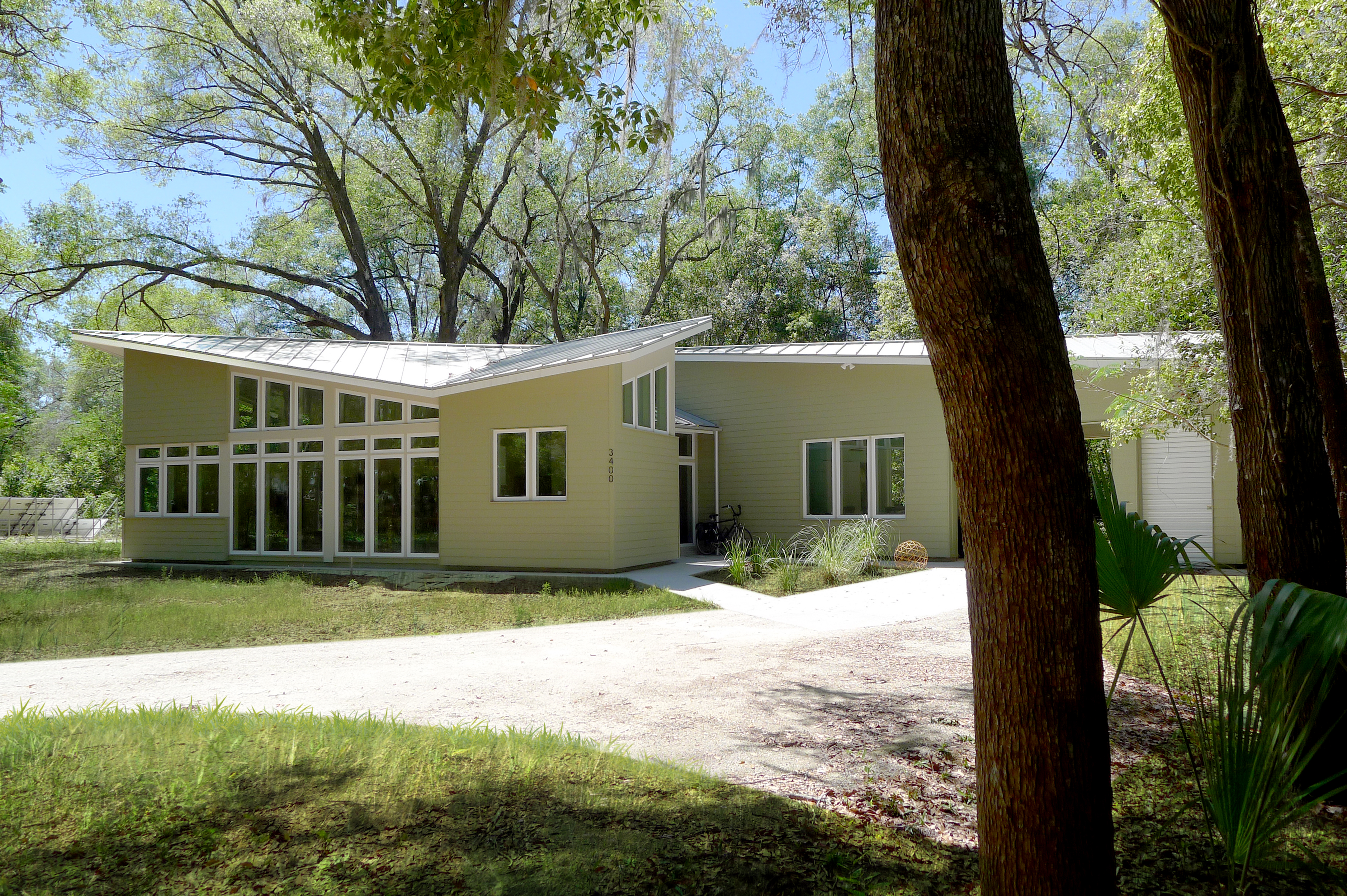 Photograph of the Quinlivan Net Zero house. The image shows a modern house, with a dramatic sloped metal roof, horizontal siding, and large glass areas. In the foreground is a driveway and trees.