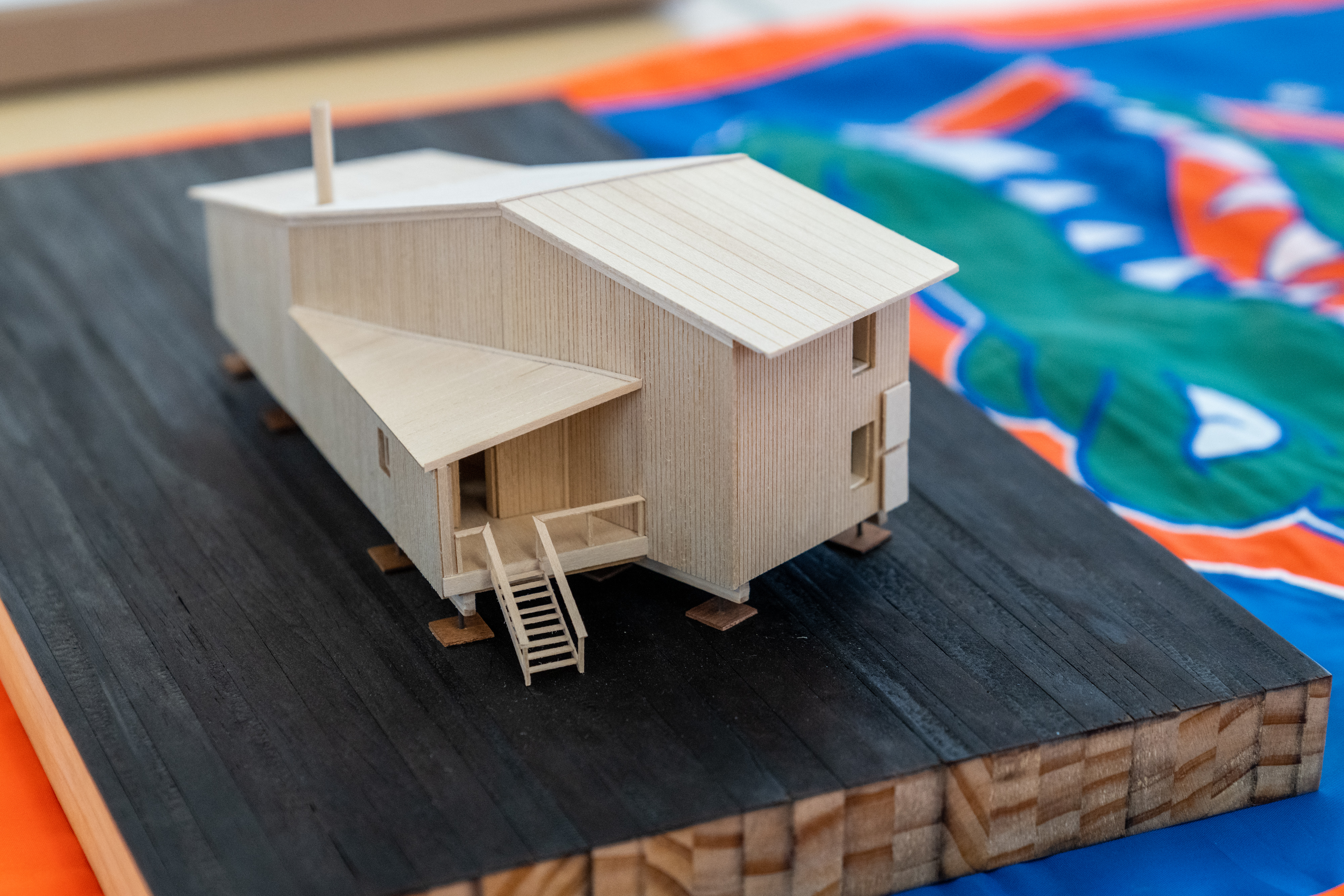 A photograph of an architectural model, built of basswood. The structure is two stories, with a simple sloping roof and separate one-story entrance. In the background, we see a banner with a logo from the University of Florida.