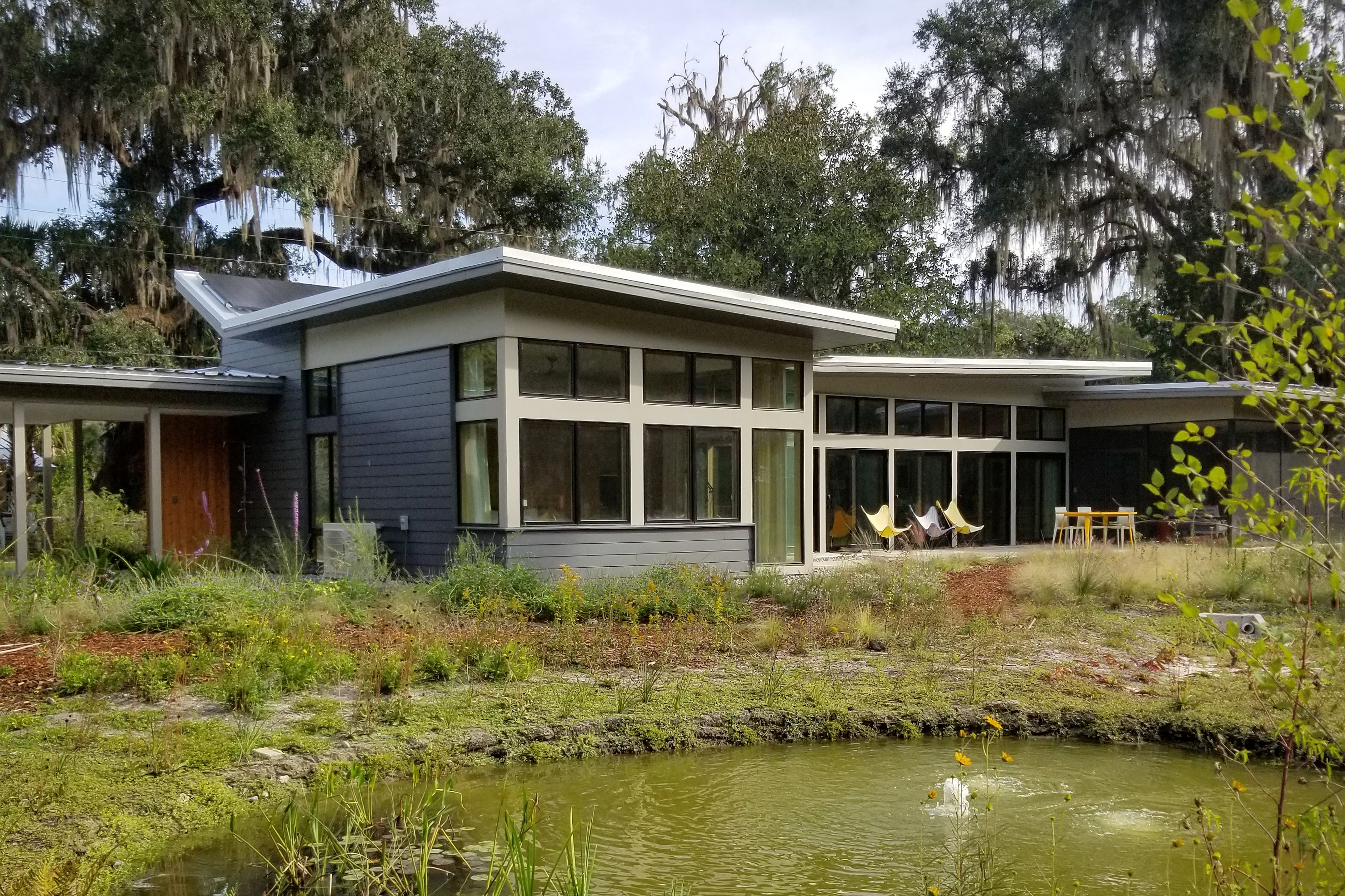 Photograph of the A2 House in Micanopy, Florida U.S.A. The image shows a one-story modern house with low-slope metal roofs. In the foreground of the image is a lush garden and pond.