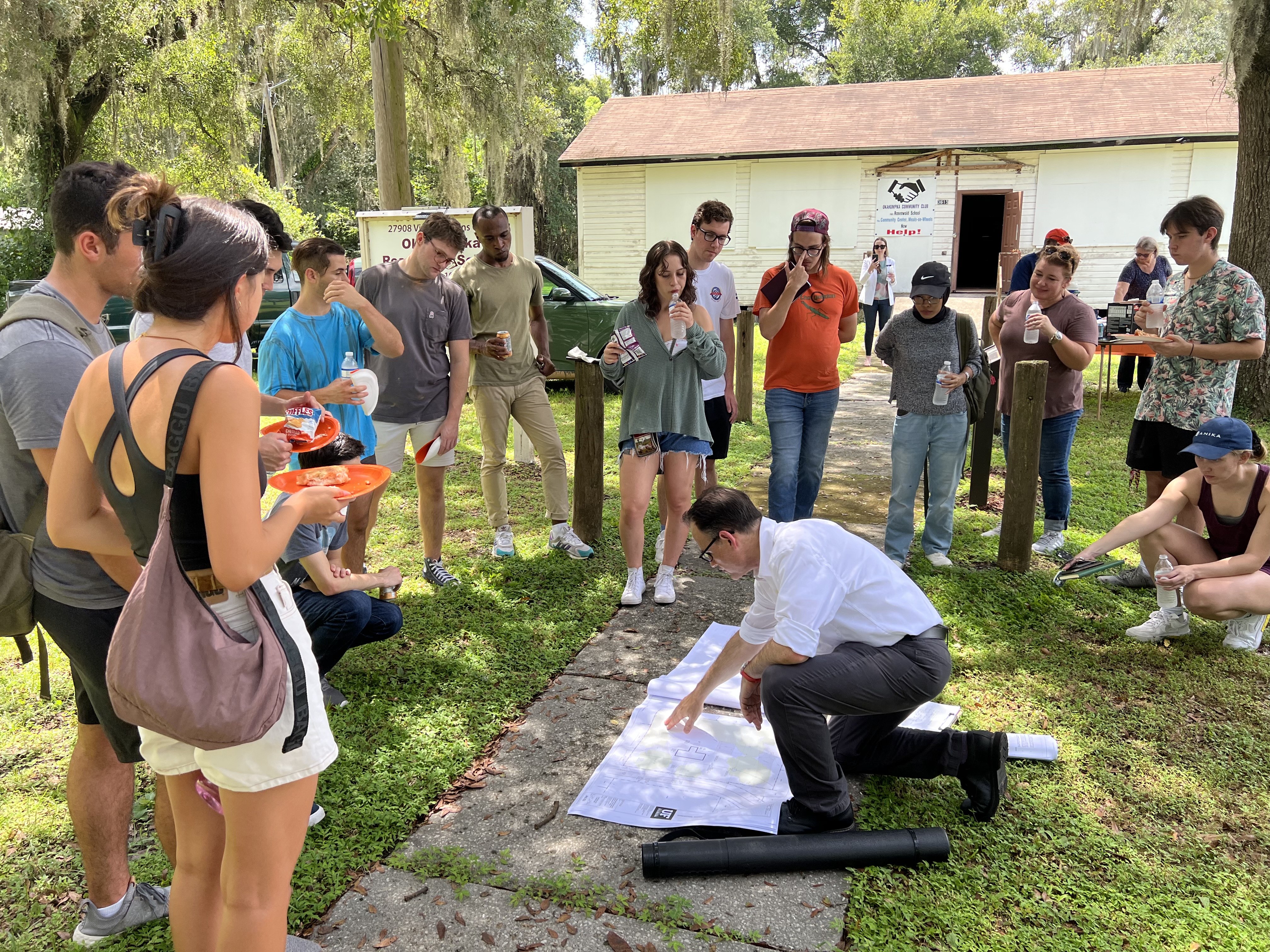 A group of students and faculty are gathered around Professor Walters, who is pointing to a large set of architectural drawings on the ground at the center of the group. In the background is the Okahumpka Rosenwald School prior to renovation, with boarded-up windows.